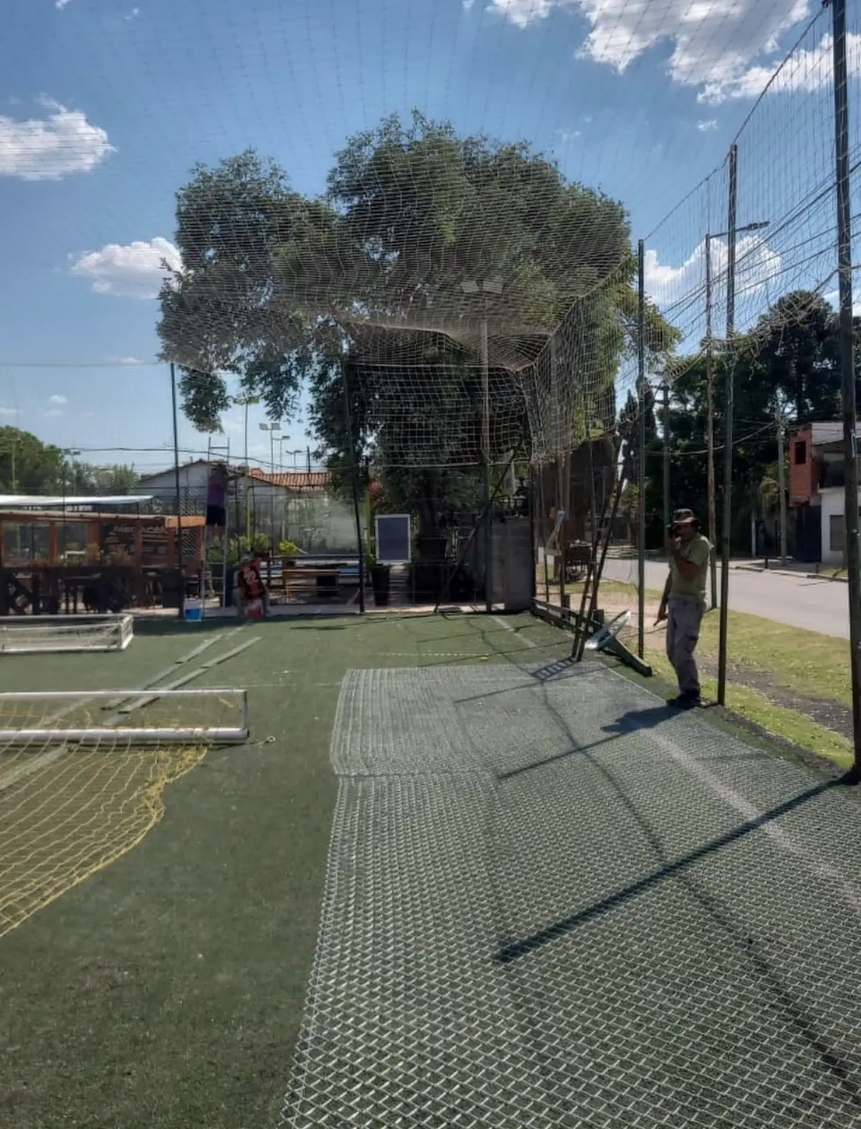 Cancha de fútbol 5 con césped sintético y arco, con el paño de tejido del alambrado perimetral apoyado en el piso durante la colocación