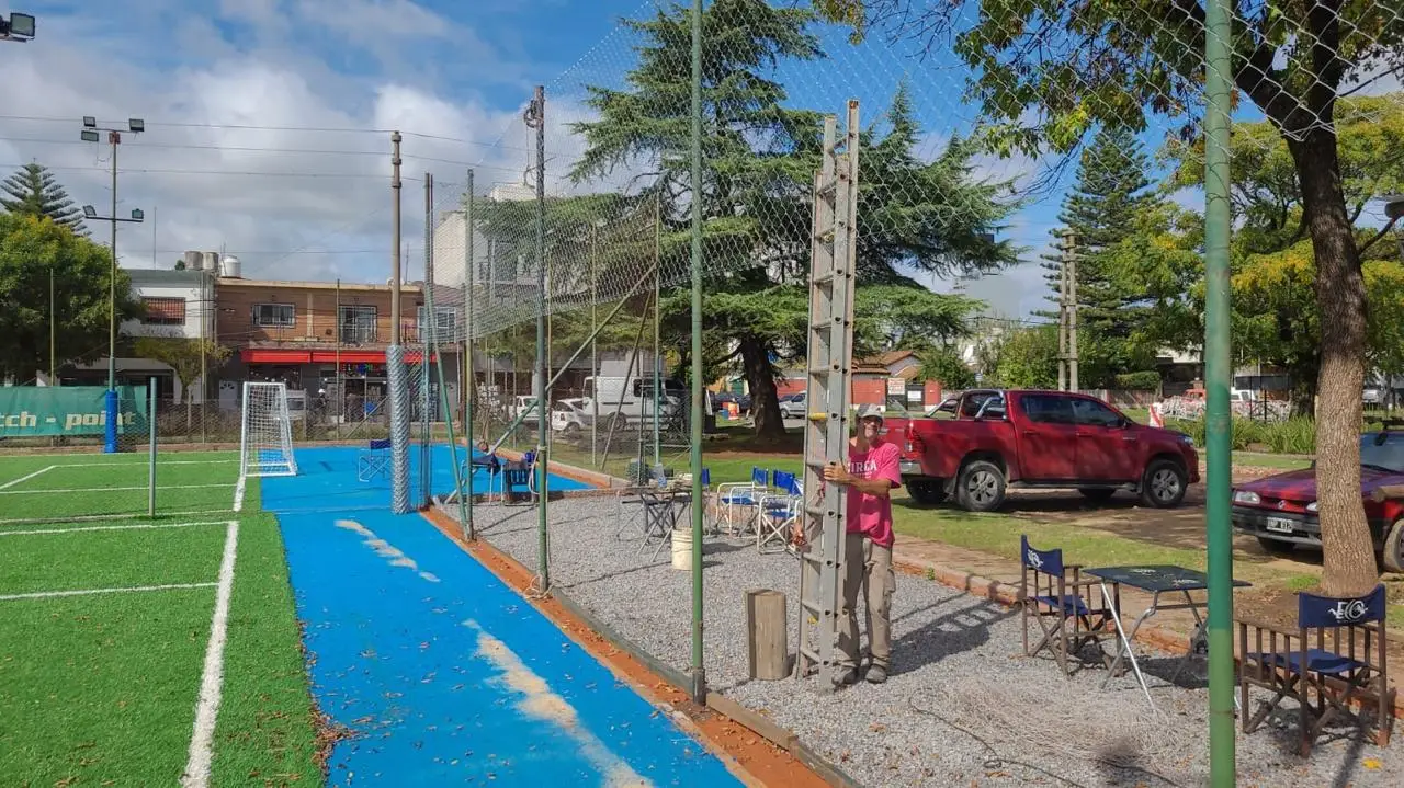 Operario de Casa Ledesma con una escalera junto al alambrado perimetral de una cancha de césped sintético con pista azul, mientras se levanta el paño entre los postes de caño