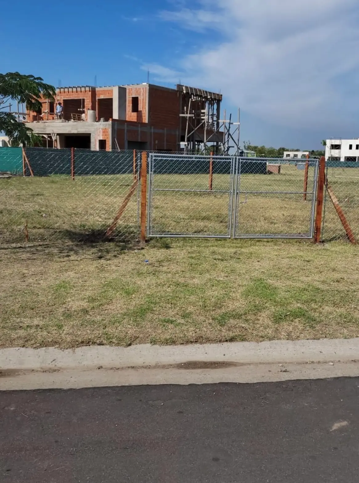 Portón de dos hojas de caño colocado en el frente de un lote, entre postes de quebracho y alambrado romboidal, con una casa en construcción atrás
