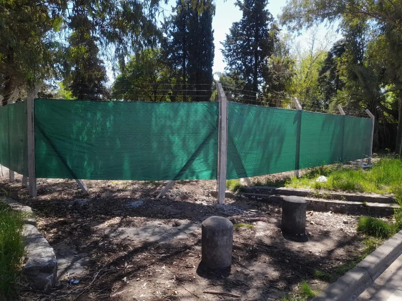 Alambrado alto con hilos tendidos en el tramo superior inclinado y media sombra verde puesta por detrás, entre árboles, con esquineros apuntalados