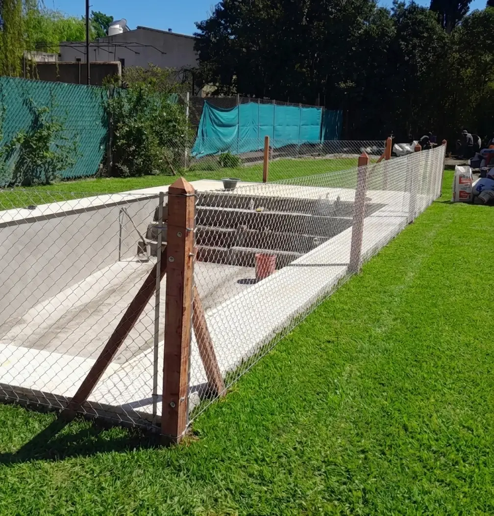 Cierre de pileta terminado: alambrado de tejido sobre postes de quebracho, con esquineros apuntalados, rodeando una pileta en un jardín con césped