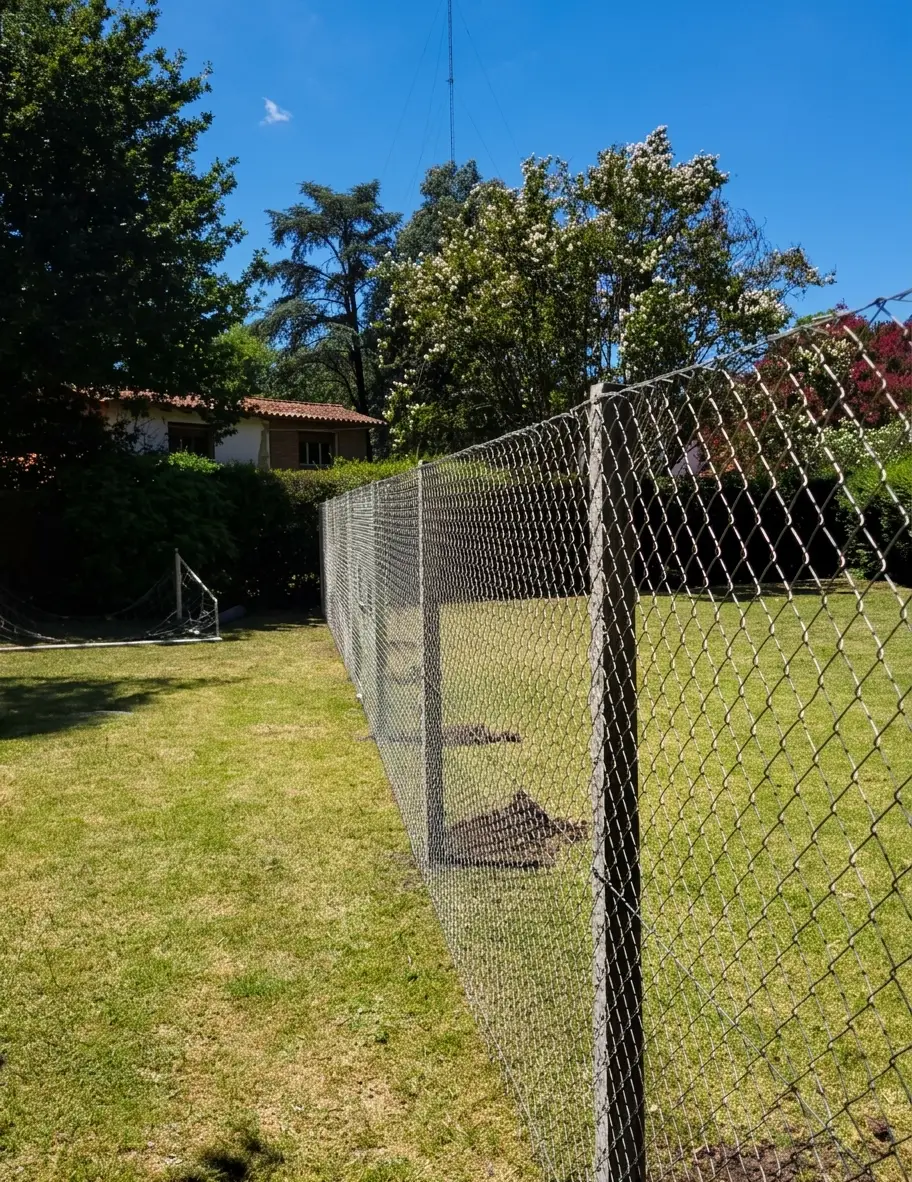 Alambrado de tejido sobre postes rectos de hormigón cerrando el fondo de un jardín con césped y árboles